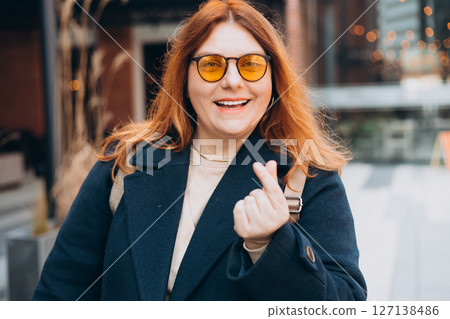 Female hand showing finger heart or mini heart gesture with the index finger and thumb come together like a snap outdoors. 30s Woman in sunglasses on urban background. High quality photo. 127138486