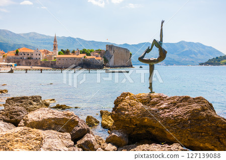 Bronze dancer statue on rocky shore with historic coastal town and mountains in background. Artistic sculpture and Mediterranean scenery. Montenegro, Budva 127139088