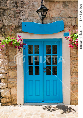 Blue wooden double door with glass panels and flowers on stone wall. Mediterranean architecture, colorful entrance, and quaint historic street. 127139105