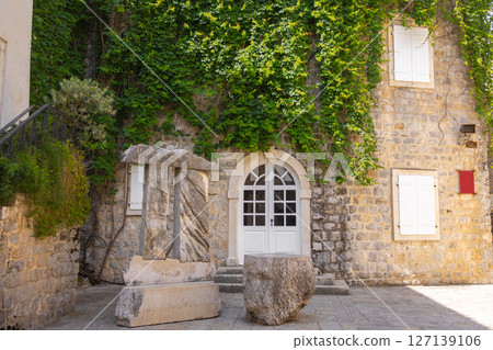 Ancient stone sculpture near ivy-covered historical building. Urban greenery, cultural heritage, and Mediterranean architecture. 127139106