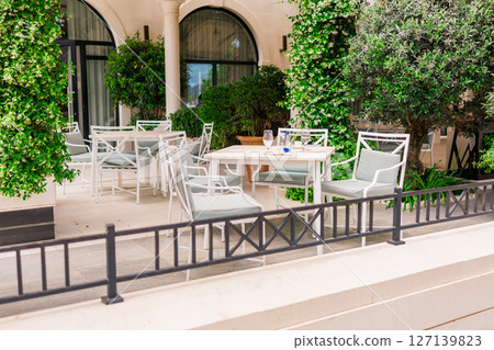 Elegant outdoor cafe terrace with white furniture and lush greenery. Relaxation, summer lifestyle, and modern hospitality. Elegant outdoor cafe terrace with white furniture and lush greenery. Relaxation, summer lifestyle, and modern hospitality. 127139823