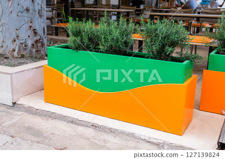 Colorful planter with herbs near outdoor cafe seating. Urban gardening, decorative design, and sustainable city landscaping. Colorful planter with herbs near outdoor cafe seating. Urban gardening, decorative design, and sustainable city landscaping. 127139824