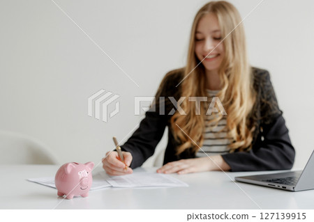 Woman writing financial documents with a piggy bank on the table in an office setting during daylight Woman writing financial documents with a piggy bank on the table in an office setting during daylight 127139915