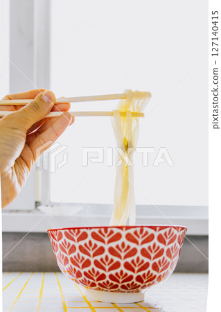 person is eating noodles with chopsticks. colorful bright red plate of ramen sits on yellow table. person is eating noodles with chopsticks. colorful bright red plate of ramen sits on yellow table. 127140415