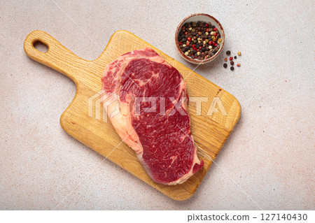 Raw ribeye steak on wooden cutting board with peppercorns top view on light beige rustic background 127140430