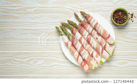 Fresh asparagus wrapped in bacon with peppercorns on white wooden table top view with space for text 127140448
