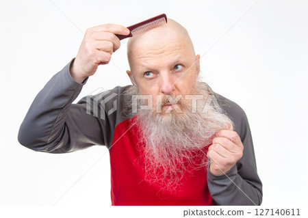 Elderly bald man with a thick white beard uses a red comb on his head, pulling beard hair  127140611