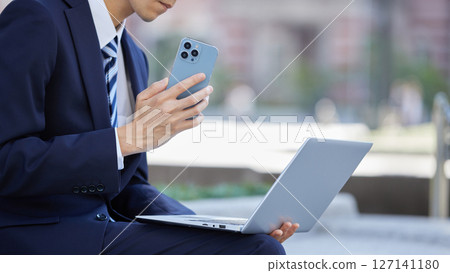 一名身著西裝的年輕男子在商業區用智慧型手機聊天 一名身著西裝的年輕男子在商業區用智慧型手機聊天 127141180