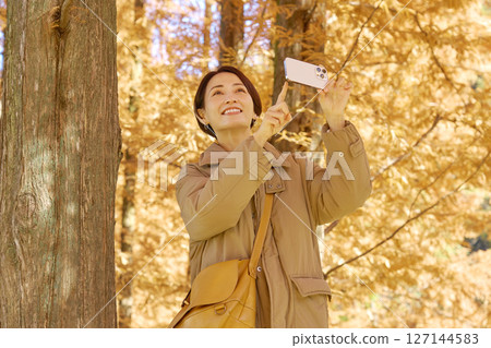 Middle-aged woman taking video of autumn leaves with her smartphone 127144583