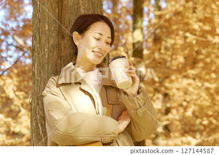 Middle-aged woman drinking coffee among autumn leaves Middle-aged woman drinking coffee among autumn leaves 127144587