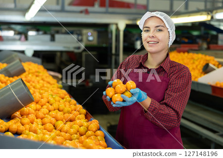 Young positive cheerful female worker in colorful uniform holds fresh ripe tangerines in her hands near the conveyor line. 127148196
