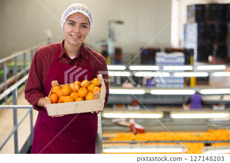 Glad positive female employee in colored uniforms hold a box of fresh ripe tangerines in their hands on citrus sorting line at warehouse. Glad positive female employee in colored uniforms hold a box of fresh ripe tangerines in their hands on citrus sorting line at warehouse. 127148203