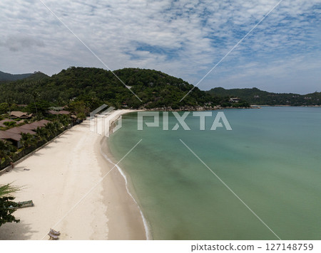 A white sand beach stretches along the turquoise water with lush green hills in the background under a partly cloudy sky. Ko Pha Ngan, Thailand. A white sand beach stretches along the turquoise water with lush green hills in the background under a partly cloudy sky. Ko Pha Ngan, Thailand. 127148759