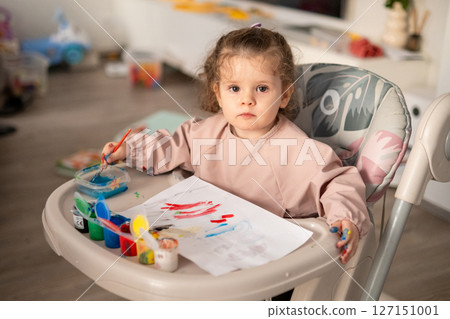 Little girl painting with gouache while sitting at a kids table. Background with soft toys and colorful childrens room decor. Creative toddler at play. 127151001