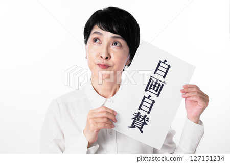 A middle-aged woman grinning and holding a piece of paper with the words "Self-praise" written on it 127151234