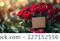 A luxurious bouquet of vibrant red roses accented with deep green foliage, tied with a crimson ribbon. A blank card is nestled within, ready for a personalized message, against a softly lit 127152556