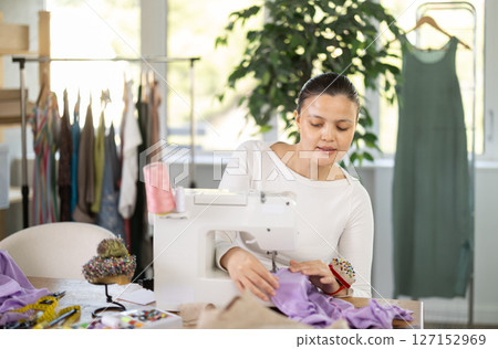 Attentive woman tailor sewing textile using sewing machine sitting at workplace in fashionable sewing atelier Attentive woman tailor sewing textile using sewing machine sitting at workplace in fashionable sewing atelier 127152969