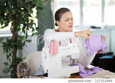 Sad young Asian tailoress viewing dress detail sitting at sewing-machine 127152999