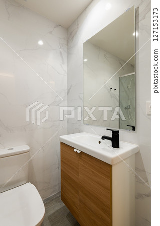 Modern bathroom corner with large mirror wood vanity white sink and black faucet against marble tile wall 127153173