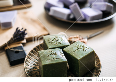 Green stamped soap bars rest on a plate beside lavender soaps and artisan tools. Artisanal process, indie skincare, microbrand identity, handmade beauty products 127154032