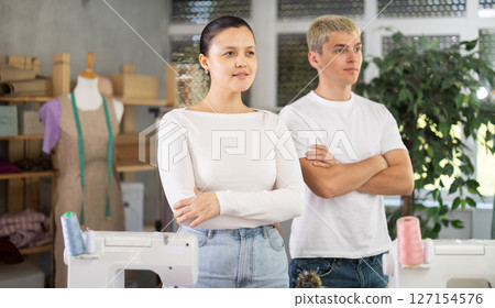 Young male and female dressmakers posing in sewing workshop Young male and female dressmakers posing in sewing workshop 127154576