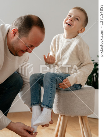 father tickles his 4-year-old son sitting on a beige chair at home. Both of European appearance are dressed in light beige clothes, the room is lit by warm sunlight with modern light interior design father tickles his 4-year-old son sitting on a beige chair at home. Both of European appearance are dressed in light beige clothes, the room is lit by warm sunlight with modern light interior design 127154855