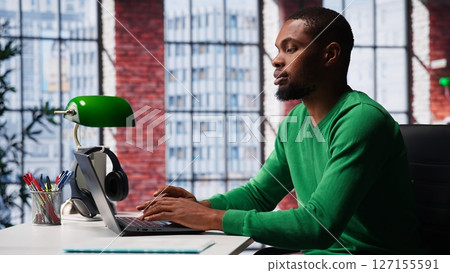 African american focused guy typing on his laptop at home office to solve daily tasks, doing remote freelance work online. Young adult surfing the web multitasking in loft apartment. Camera A. 127155591