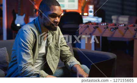 Focused African American guy teleworking on his laptop in a stylish living room. Remote worker balancing comfort and work efficiency in a modern workspace. Comfortable loft studio. Camera B. Focused African American guy teleworking on his laptop in a stylish living room. Remote worker balancing comfort and work efficiency in a modern workspace. Comfortable loft studio. Camera B. 127155605