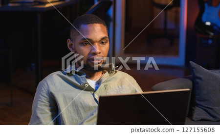 Black male freelancer taking online course to learn new skills from home, attend online video call and watching lecture from loft. Young man participates at a virtual training class. Camera B. Black male freelancer taking online course to learn new skills from home, attend online video call and watching lecture from loft. Young man participates at a virtual training class. Camera B. 127155608