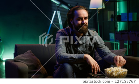 Man enjoying bowl of popcorn and smiling while watching cable programs on TV during leisure time at home. Spectator on couch in living room eating snacks and looking at television series, camera A Man enjoying bowl of popcorn and smiling while watching cable programs on TV during leisure time at home. Spectator on couch in living room eating snacks and looking at television series, camera A 127155767