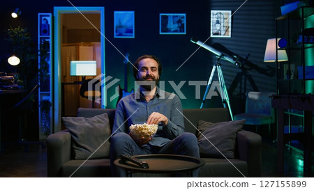 Amused person sitting on couch using TV to binge comedy series on subscription based streaming services, enjoying popcorn. Man laughing at funny shows on television, eating snack, camera B Amused person sitting on couch using TV to binge comedy series on subscription based streaming services, enjoying popcorn. Man laughing at funny shows on television, eating snack, camera B 127155899