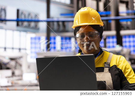 Closeup of female compliance engineer reviewing machinery software updates on her laptop. African american industrial technician working on developing innovative infrastructures in smart factory. 127155903