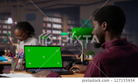 College student looks at a copy space on laptop while collecting notes from reliable academic sources, consulting the campus library books collection for an assignment. Student life. Camera A. 127155934
