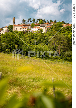 Scenic Hilltop Town Of Stanjel In Slovenia: Traditional Stone Houses And Historic Church Tower Rising Above Lush Greenery Under A Dramatic Cloudy Sky In The Karst Region 127156871