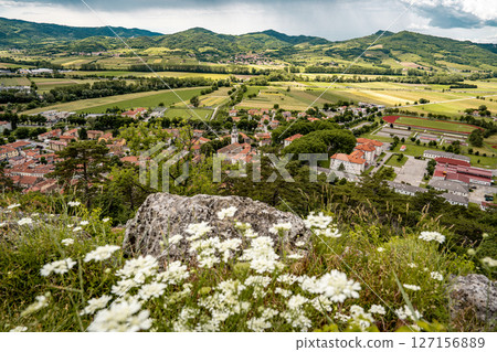 Picturesque View Of Vipava From Castle Ruins: Red-Tiled Rooftops And Church Tower Nestled In Green Valley With Blooming Flowers In Foreground And Rolling Fields In Background, Slovenia 127156889