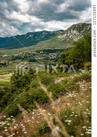 Mountain View Toward Ajdovscina From Vipava Castle: Rolling Green Hills, Wildflowers, And A Distant Town Nestled Below Steep Rocky Peaks Under A Moody Summer Sky In Western Slovenia 127156895