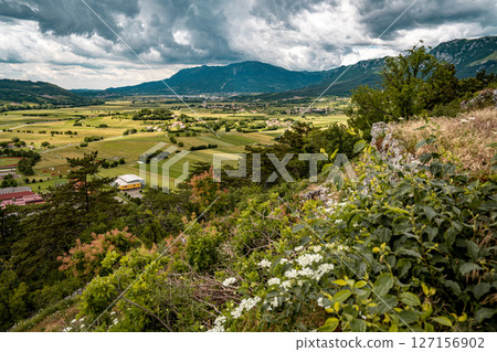 Mountain View Toward Ajdovscina From Vipava Castle: Rolling Green Hills, Wildflowers, And A Distant Town Nestled Below Steep Rocky Peaks Under A Moody Summer Sky In Western Slovenia Mountain View Toward Ajdovscina From Vipava Castle: Rolling Green Hills, Wildflowers, And A Distant Town Nestled Below Steep Rocky Peaks Under A Moody Summer Sky In Western Slovenia 127156902