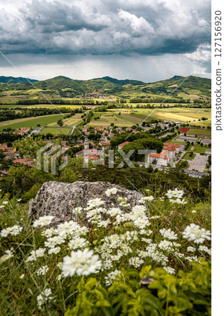 Picturesque View Of Vipava From Castle Ruins: Red-Tiled Rooftops And Church Tower Nestled In Green Valley With Blooming Flowers In Foreground And Rolling Fields In Background, Slovenia Picturesque View Of Vipava From Castle Ruins: Red-Tiled Rooftops And Church Tower Nestled In Green Valley With Blooming Flowers In Foreground And Rolling Fields In Background, Slovenia 127156920