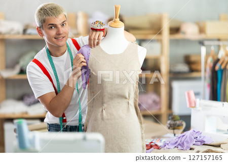 Experienced young male fashion designer working with fabric and mannequin while sewing fashionable dress in sewing workshop Experienced young male fashion designer working with fabric and mannequin while sewing fashionable dress in sewing workshop 127157165