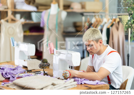 Professional young attentive man seamstress sewing textile using sewing machine sitting at table at workplace in sewing studio 127157265