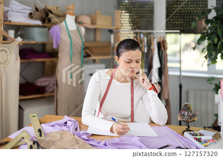 Frustrated young female dressmaker reading clothing manufacturing documentation or paper report finding problems in order calculations while working in sewing workshop 127157320