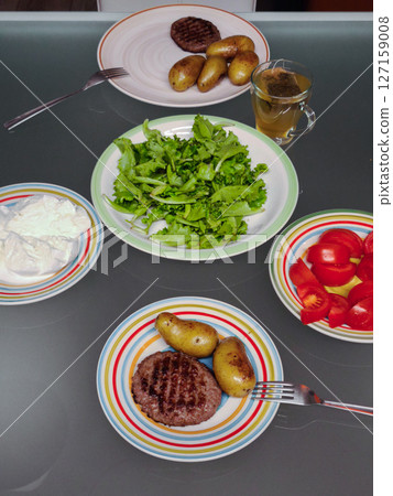 Grilled raw burger patties beside boiled potatoes, green salad, tomato slices, herbal tea on glass table 127159008