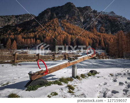 Wooden seesaw sitting on snow dusted ground, encircled by golden trees and alpine peaks in aosta valley, winter approaching Frazionne Lillaz Wooden seesaw sitting on snow dusted ground, encircled by golden trees and alpine peaks in aosta valley, winter approaching Frazionne Lillaz 127159037