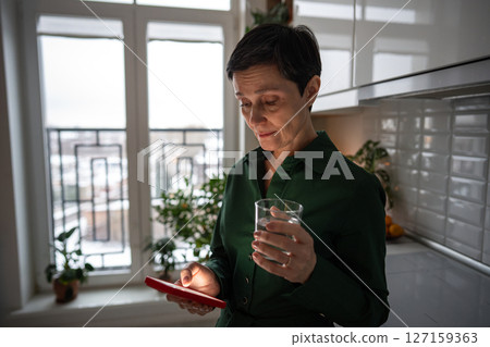 Serious woman staring at smartphone screen with water glass in kitchen, distress. Bad news stress 127159363