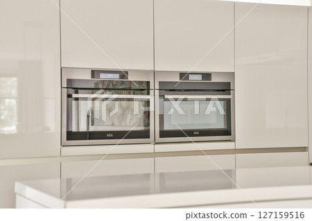 A sleek, contemporary kitchen showcases built-in ovens with a minimalist design. Smooth surfaces and neutral colors create a sophisticated atmosphere. 127159516