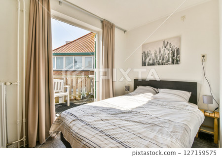 A cozy modern bedroom featuring a neatly made bed, large window with natural light, and a view of an outdoor balcony with seating. 127159575