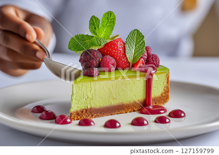 Chef carefully plates a vibrant pistachio cheesecake slice adorned with berries and coulis in an upscale restaurant setting. Bright colors enhance presentation, emphasizing culinary artistry 127159799