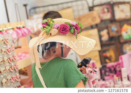 Woman in wide straw hat adorned with vivid red and pink roses at outdoor market. Concept of summer fashion, summer accessories, summer vibe Woman in wide straw hat adorned with vivid red and pink roses at outdoor market. Concept of summer fashion, summer accessories, summer vibe 127161578