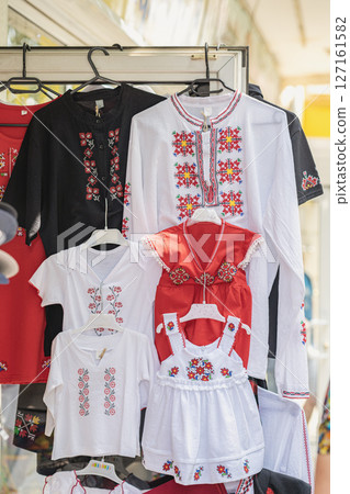 Embroidered traditional national bulgarian shirts and folk dresses for adults and children hanging at colorful market stall. Concept of family heritage, ethnic clothing, cultural identity Embroidered traditional national bulgarian shirts and folk dresses for adults and children hanging at colorful market stall. Concept of family heritage, ethnic clothing, cultural identity 127161582