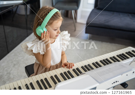 Little girl practicing piano with wireless headphones at home. Early childhood development through modern and focused music education. Little girl practicing piano with wireless headphones at home. Early childhood development through modern and focused music education. 127161965